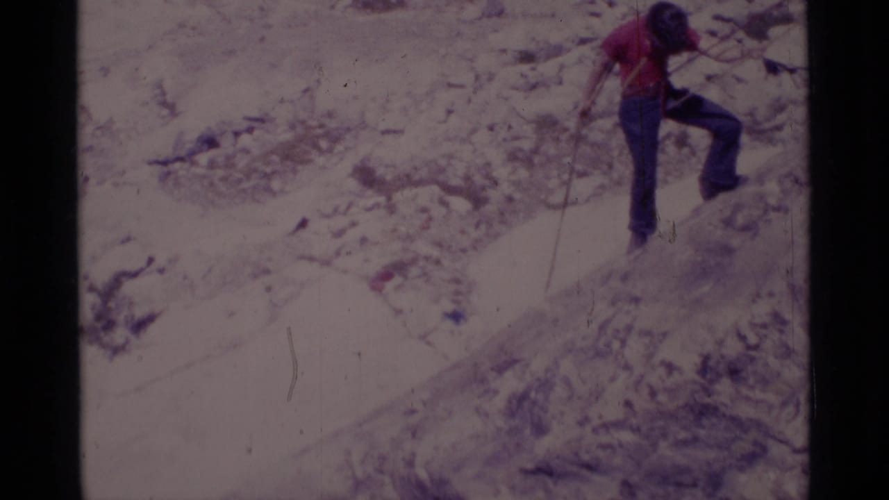 The red-shirted climber continues moving across a steep rock face, with rope visible against the pale terrain.