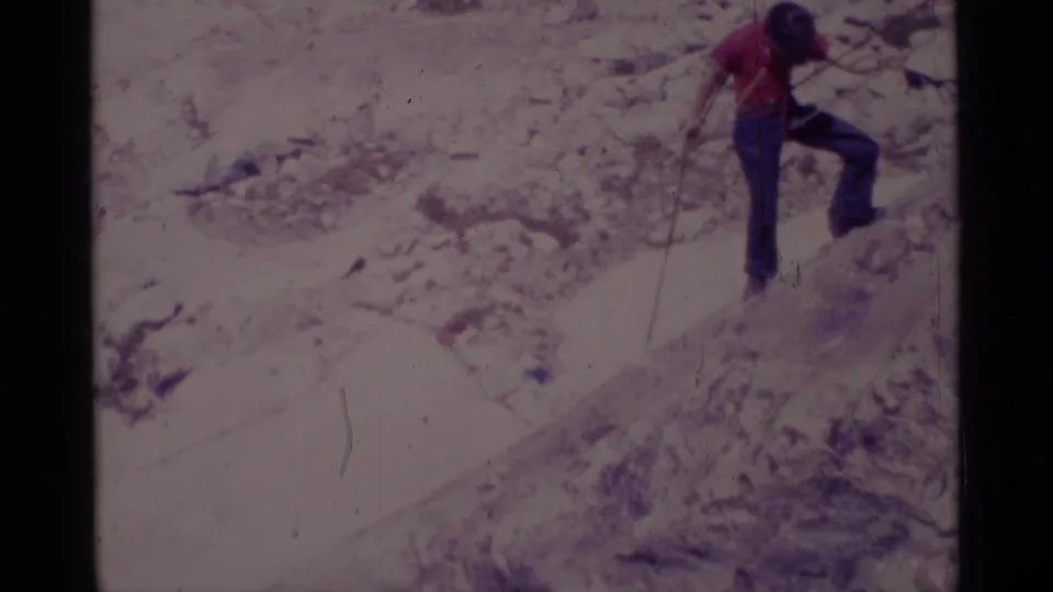 The red-shirted climber continues moving across a steep rock face, with rope visible against the pale terrain.