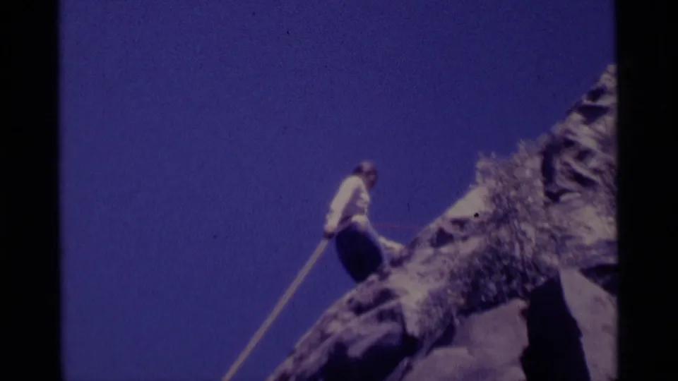 A climber in a white shirt moves on a rocky outcrop with a rope visible, framed against deep blue sky.