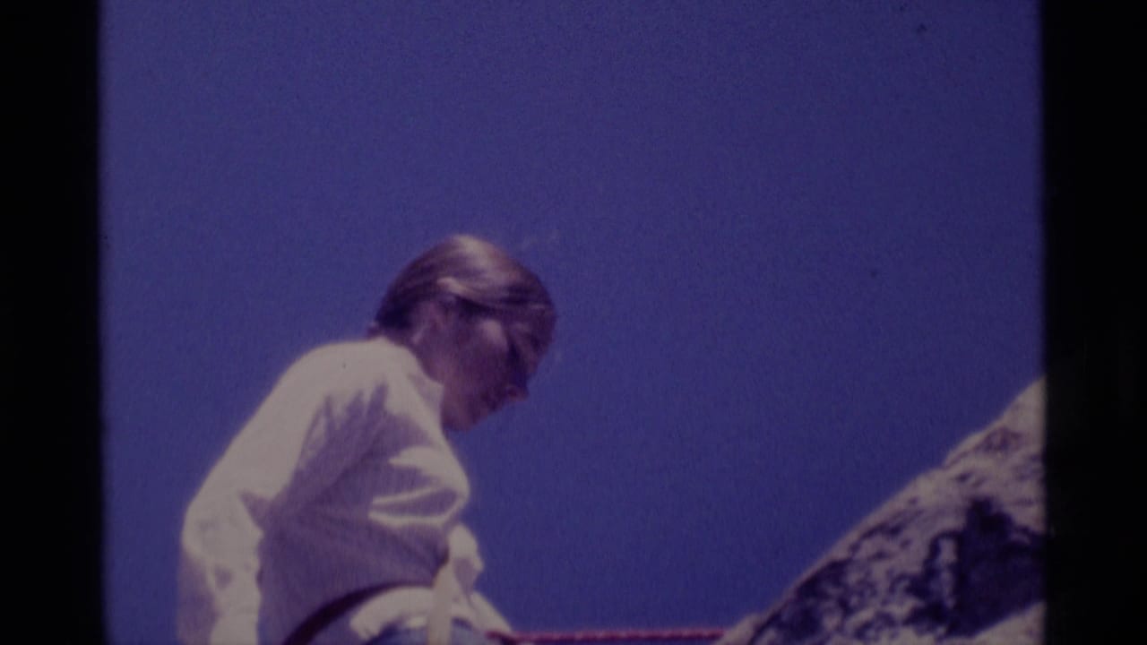 1975 8mm frame from Minaret Wilderness showing a woman in a light shirt beside a rock face with rope visible against blue sky.