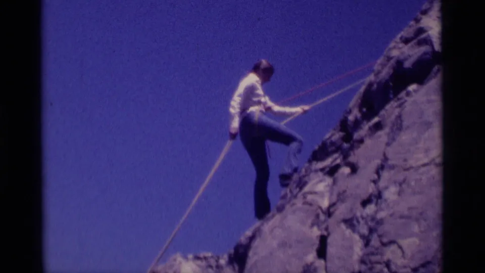 1975 8mm frame from Minaret Wilderness showing a woman descending a steep rock face on rope beneath a clear blue sky.