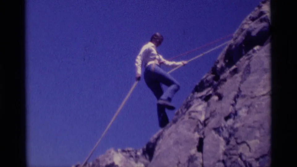 1975 8mm frame showing a woman in Minaret Wilderness rappelling partway down a rough gray cliff face.