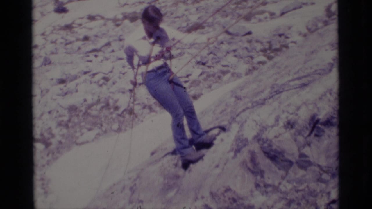 Roped climber in jeans and a light shirt pauses on sloping rock in the California wilderness footage.