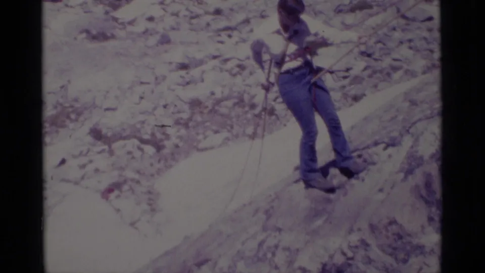 Home-movie frame of a climber in jeans and a light shirt balancing with ropes on rocky wilderness terrain.