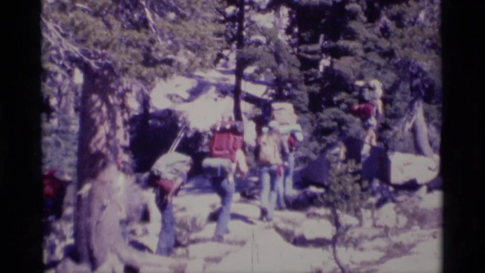 1975 Minaret Wilderness frame showing backpackers in line on a rocky trail through dense trees.