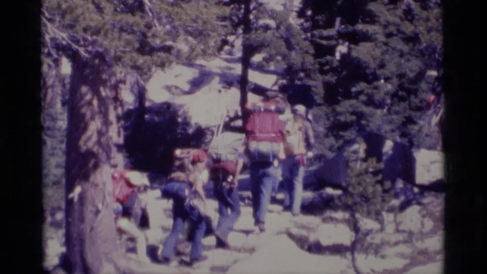 1975 California wilderness home-movie frame with backpackers moving away along a rocky forest trail.