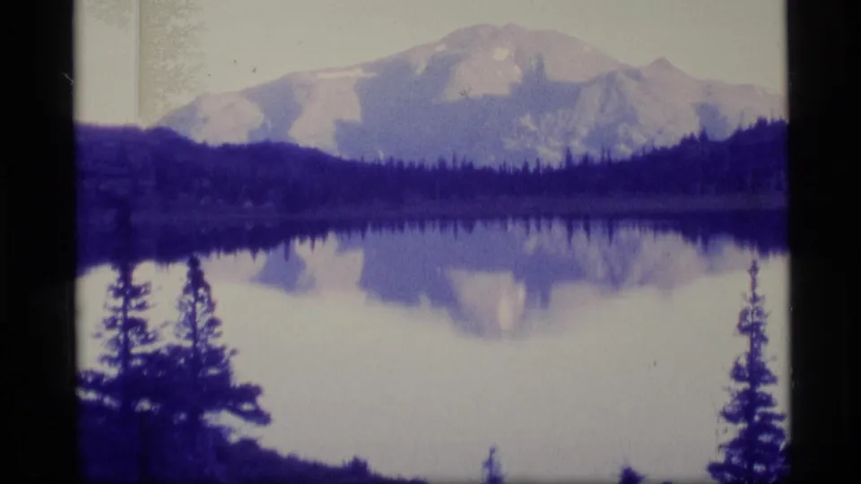 Wide 1977 Alaska scenic view of a mountain reflected in a still lake, framed by dark evergreen silhouettes.