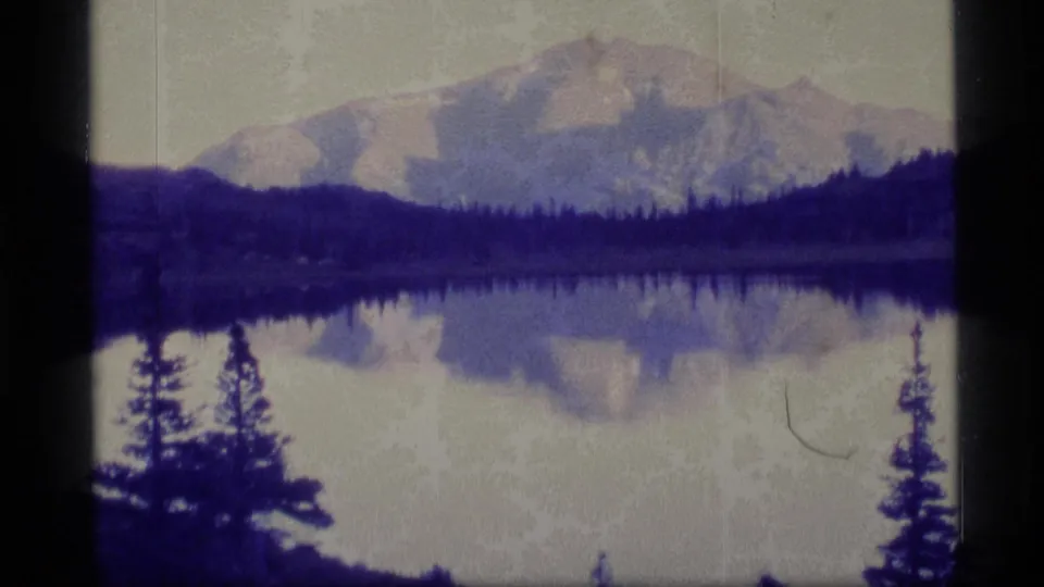 Late 1970s Alaska home-movie frame of a mountain reflected across a quiet lake, with evergreens and aged film texture.