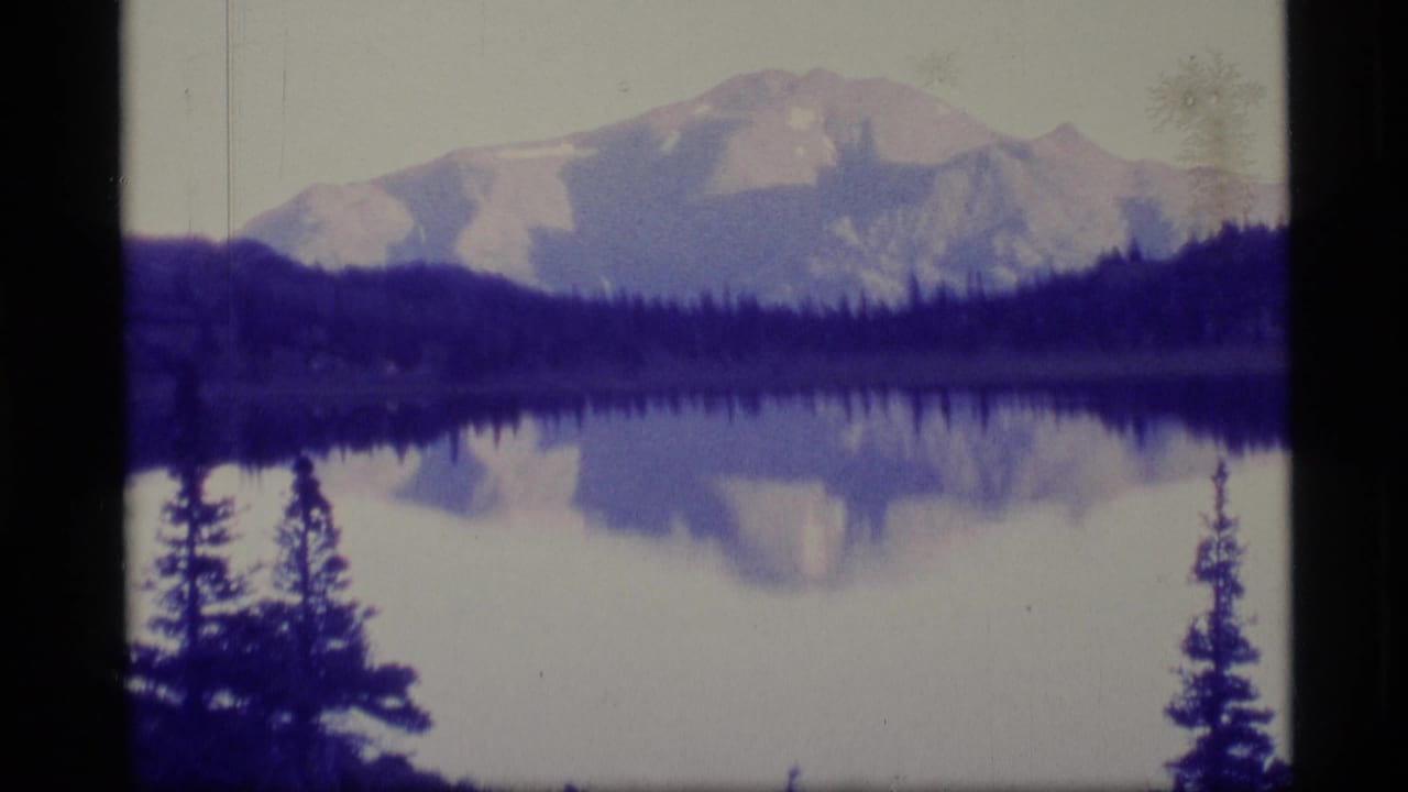 1977 Alaska 8mm frame showing a mountain reflected in a still lake, with dark evergreen trees framing the shoreline.