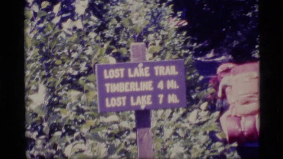1977 Alaska 8mm frame with a Lost Lake Trail sign listing Timberline and Lost Lake distances, beside a partly visible backpacked hiker.