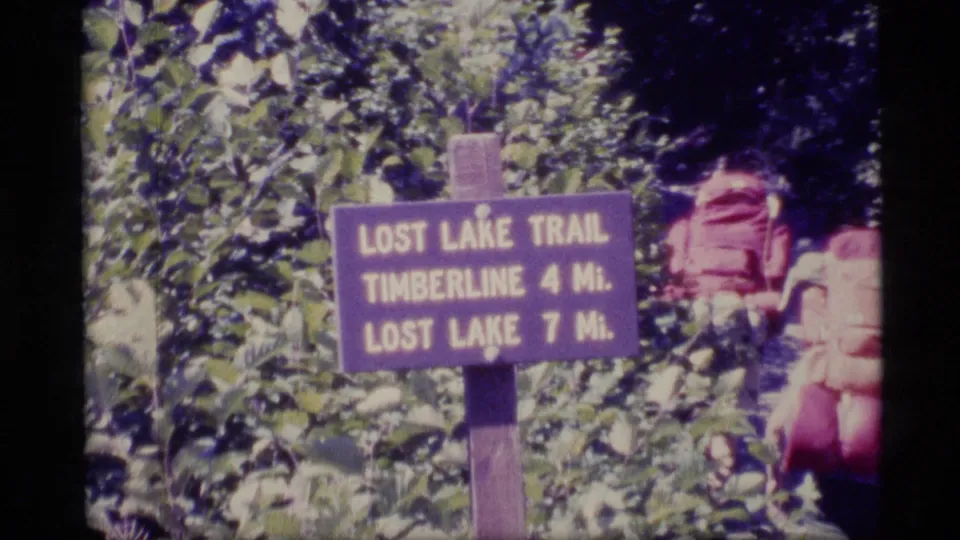 1977 Alaska 8mm frame showing a Lost Lake Trail sign in leafy brush, with backpacking gear and a hiker partly visible at right.