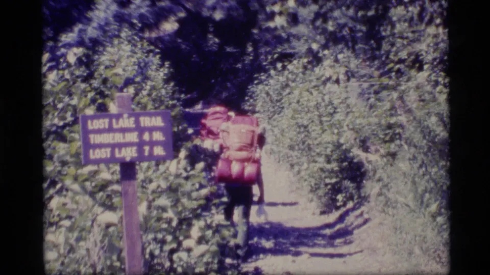 1977 Alaska 8mm frame showing a hiker with a large pack walking down a forest trail past the Lost Lake Trail sign.