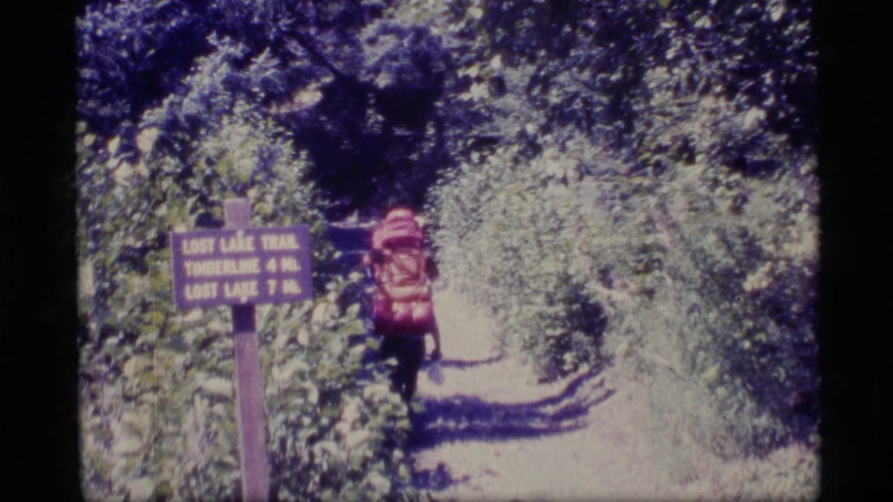 1977 Alaska 8mm frame of a hiker with a red rucksack walking away past a wooden Lost Lake Trail sign in dense greenery.