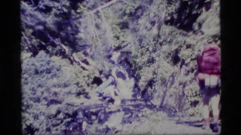 1977 Alaska home-movie frame showing a backpacker in a red pack walking through dense forest.