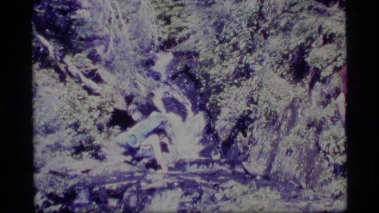 1977 Alaska 8mm frame showing a backpacked hiker crouched near fast water among rocks and greenery.