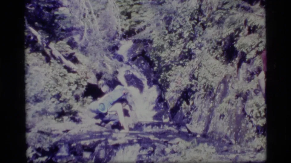 1977 Alaska 8mm frame showing a backpacked hiker crouched near fast water among rocks and greenery.