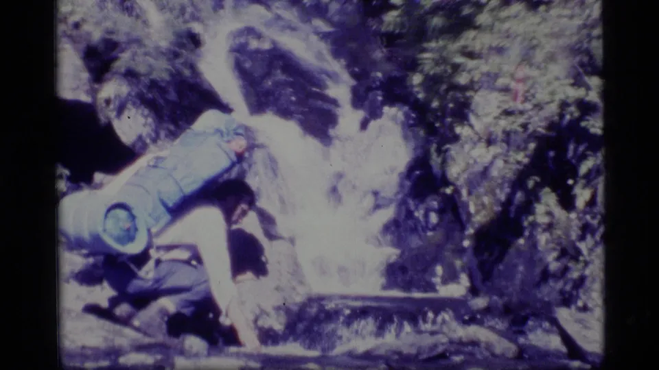 Close 1977 Alaska home-movie view of a hiker with a blue pack collecting or reaching toward mountain stream water.