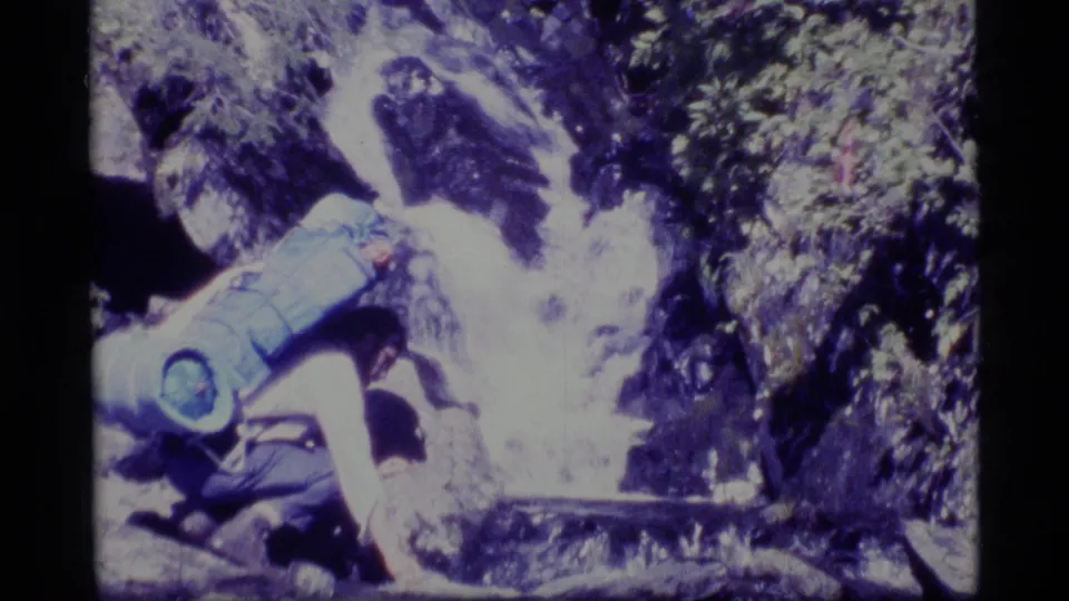 1977 Alaska 8mm still of a backpacked hiker crouched at a rocky mountain stream in dense greenery.