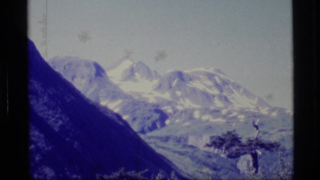 1977 Alaska 8mm frame showing distant snow-capped mountains and a dark foreground valley slope beneath a pale sky.