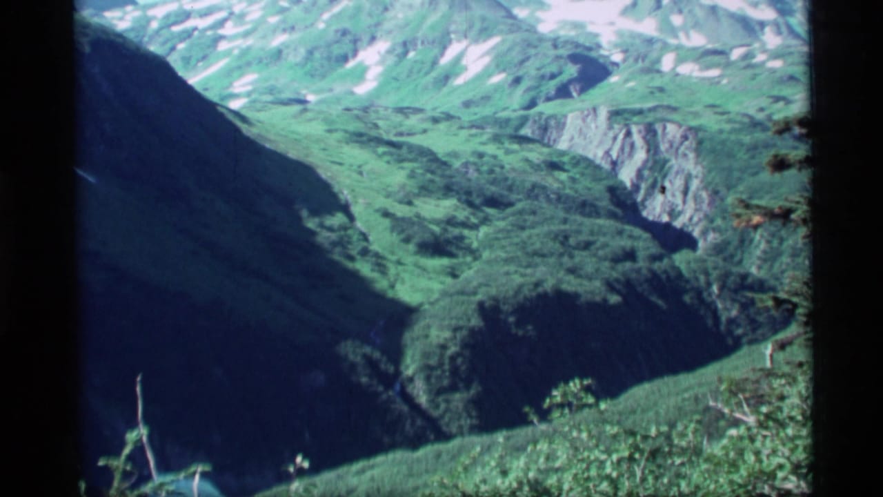Green mountain valley with steep slopes and snow patches in the distance.