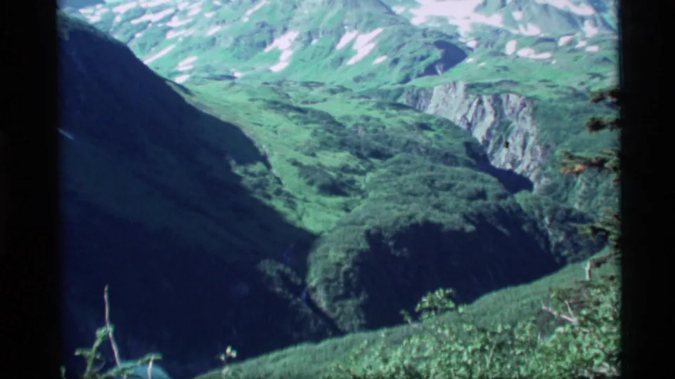 1977 Alaska 8mm view across a green mountain valley with steep shadowed slopes and snow-dusted peaks beyond.