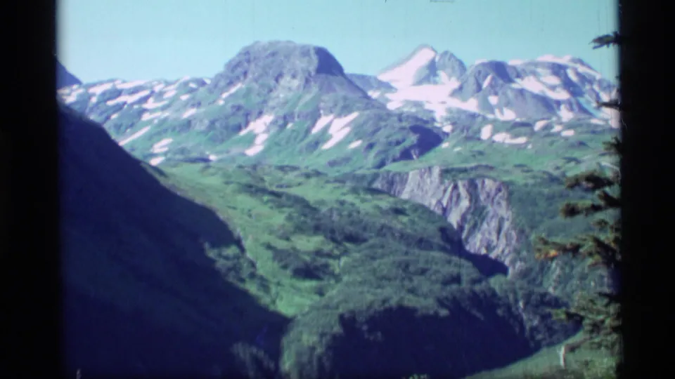 1977 Alaska 8mm panoramic frame of rugged mountains, green slopes, rocky faces, and lingering snow under pale blue sky.
