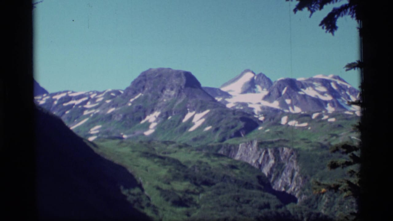 1977 Alaska 8mm frame showing distant snow-capped mountains, green ridges, and dark tree edges under clear sky.