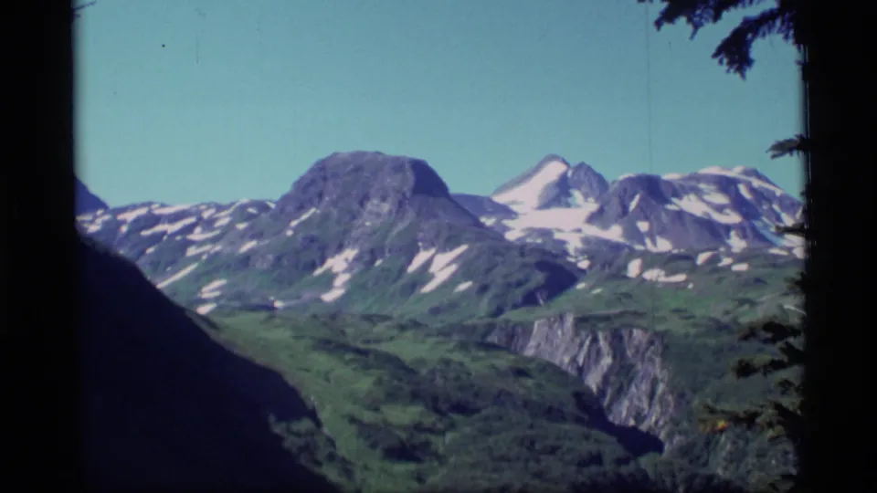 1977 Alaska 8mm frame showing distant snow-capped mountains, green ridges, and dark tree edges under clear sky.