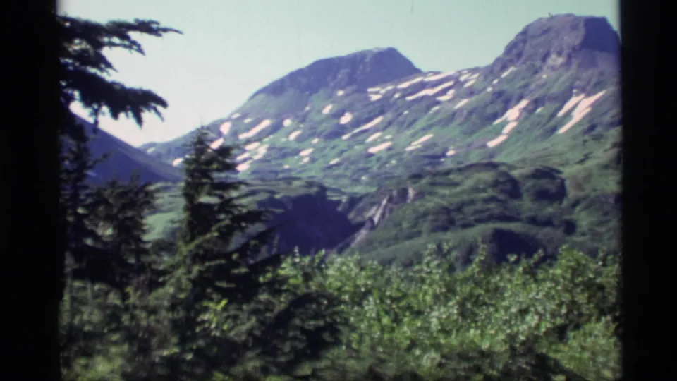 1977 Alaska home-movie view of snow-patched mountain peaks rising beyond green slopes and foreground evergreens.