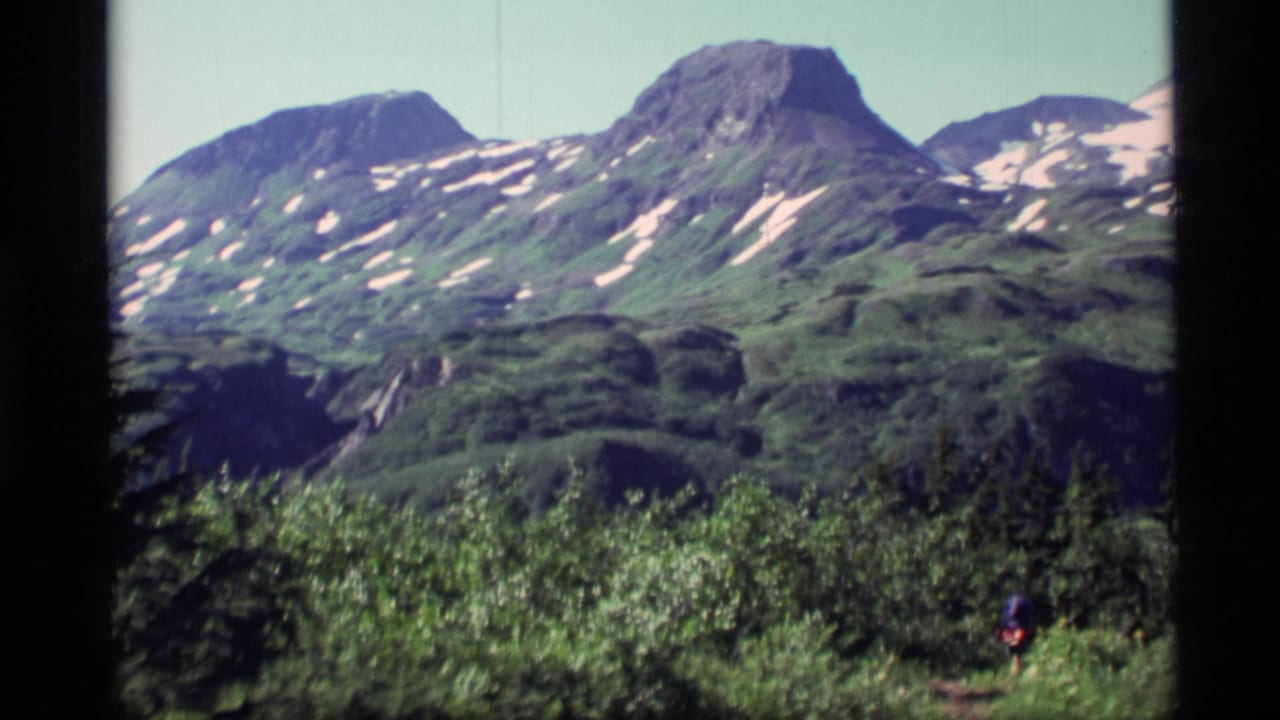 1977 Alaska 8mm frame showing green mountain slopes with snow patches, brushy foreground, and a distant hiker on a path.
