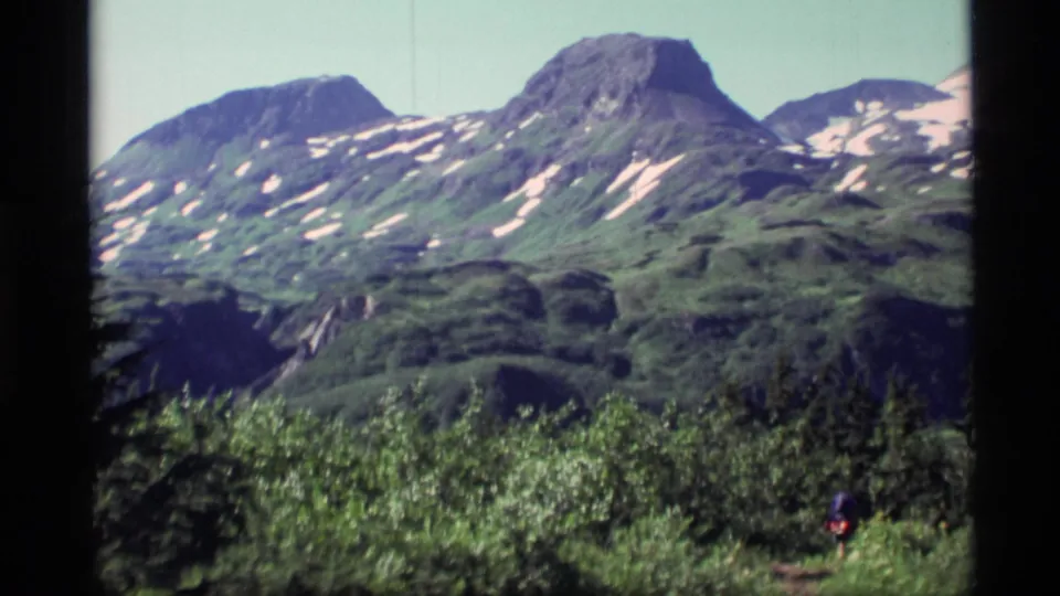 1977 Alaska 8mm frame showing green mountain slopes with snow patches, brushy foreground, and a distant hiker on a path.