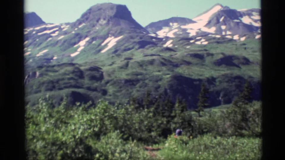 1977 Alaska home-movie frame with snow-streaked peaks, green valley ridges, dense brush, and a small hiker below.