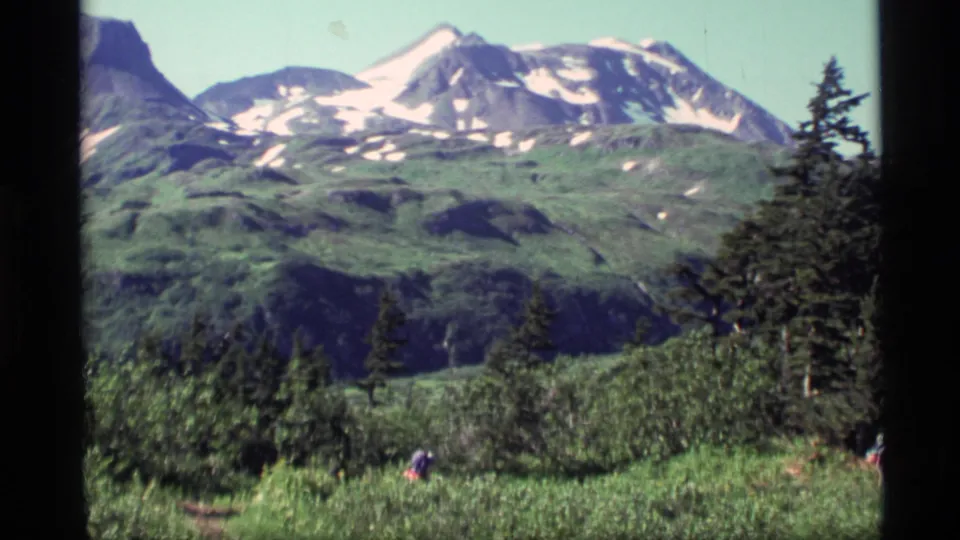 1977 Alaska frame showing a green valley below snow-patched mountains, with evergreen trees and a distant hiker in the foreground.
