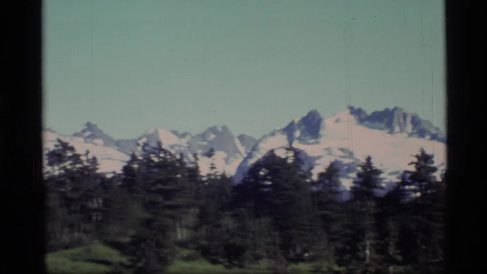 1977 Alaska 8mm frame with jagged snow-covered peaks, dark tree line, green foreground, and visible film wear.
