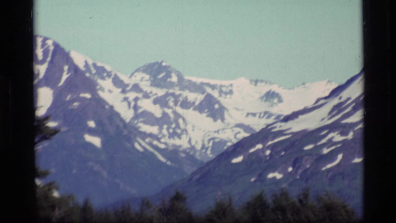 1977 Alaska home-movie frame showing distant snowy mountains above a dark forested foreground and pale blue sky.