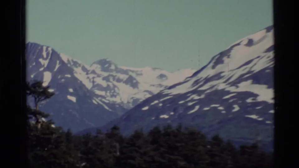 1977 Alaska landscape frame with layered blue mountain ridges, snow patches and evergreen forest in the foreground.