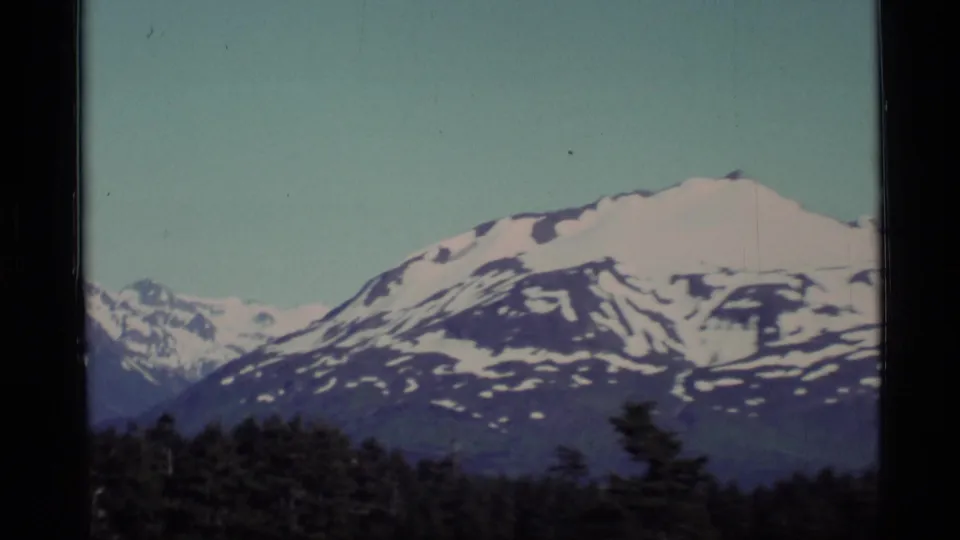 1977 Alaska 8mm frame showing a broad snow-streaked mountain slope with forest and distant peaks below.