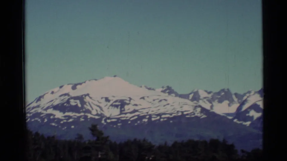 1977 Alaska home-movie frame with a snowy ridge, jagged distant peaks and dark evergreen treetops below.
