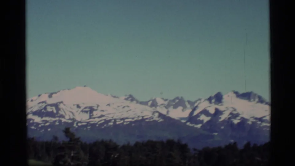 1977 Alaska 8mm frame showing snowy mountain peaks above a dark evergreen foreground and pale blue sky.