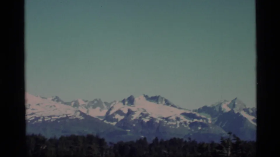 1977 Alaska 8mm view of a broad snowy mountain range seen across a dark tree line.