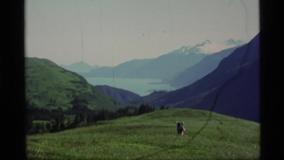 1977 Alaska 8mm frame showing a distant hiker in a green alpine meadow with mountains and possible water beyond.