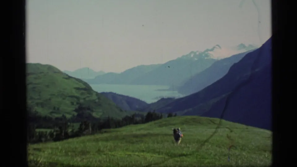 A distant hiker with a backpack crosses an Alaska alpine plateau, framed by green hills, lake water, and a layered mountain range.