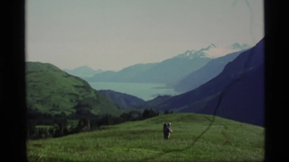 1977 home-movie still of a backpacker traversing a grassy Alaska plateau above a distant lake and layered mountains.