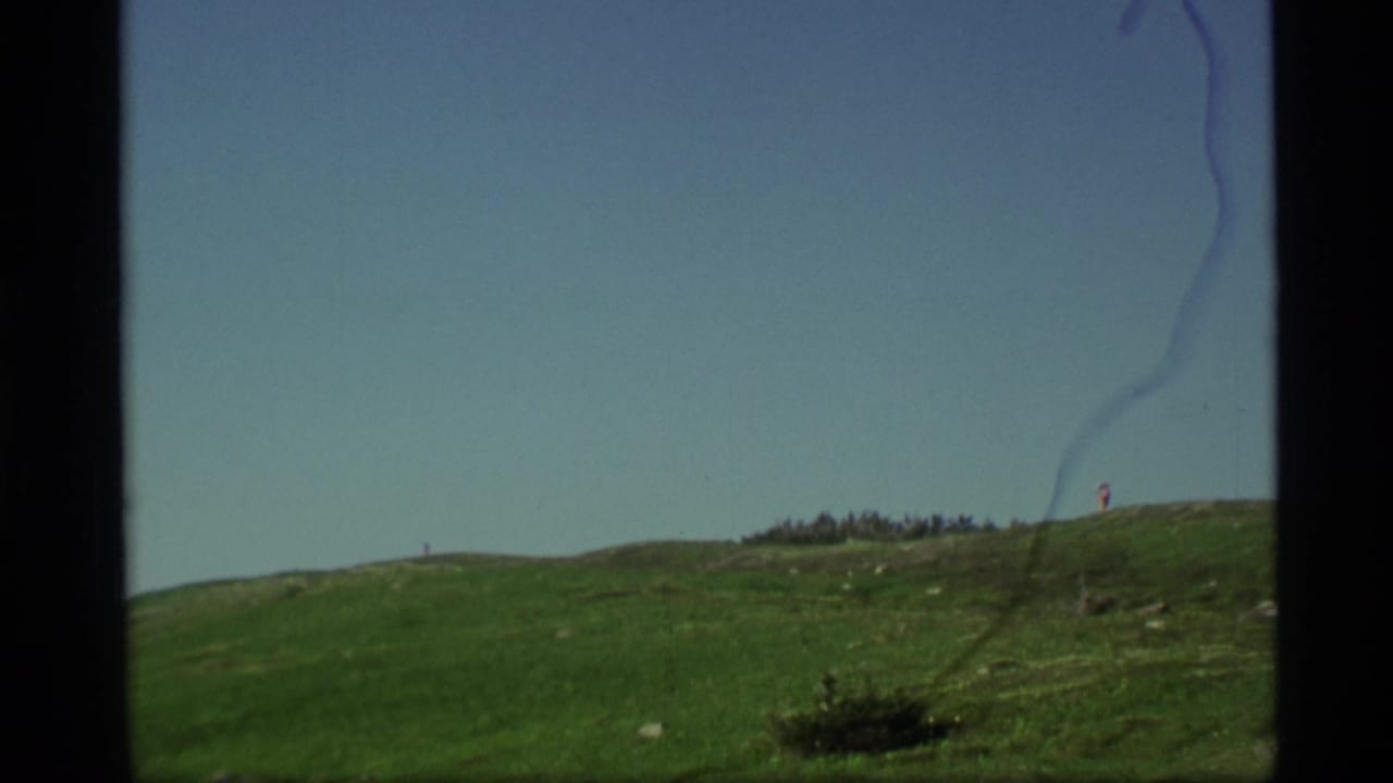 1977 Alaska 8mm frame showing a green hillside, blue sky, and a distant hiker or figure near the ridge line.