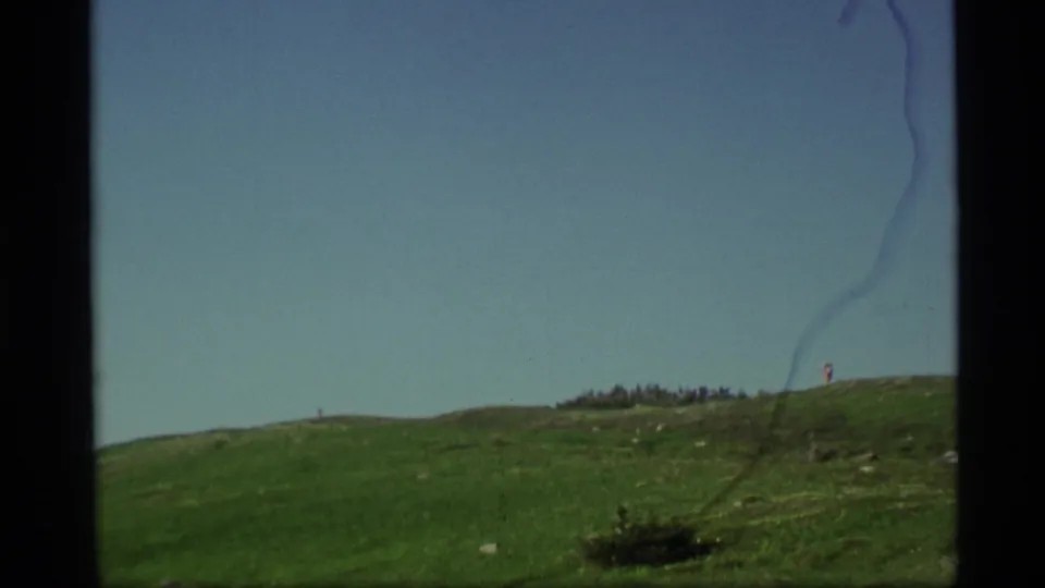 1977 Alaska 8mm frame showing a green hillside, blue sky, and a distant hiker or figure near the ridge line.