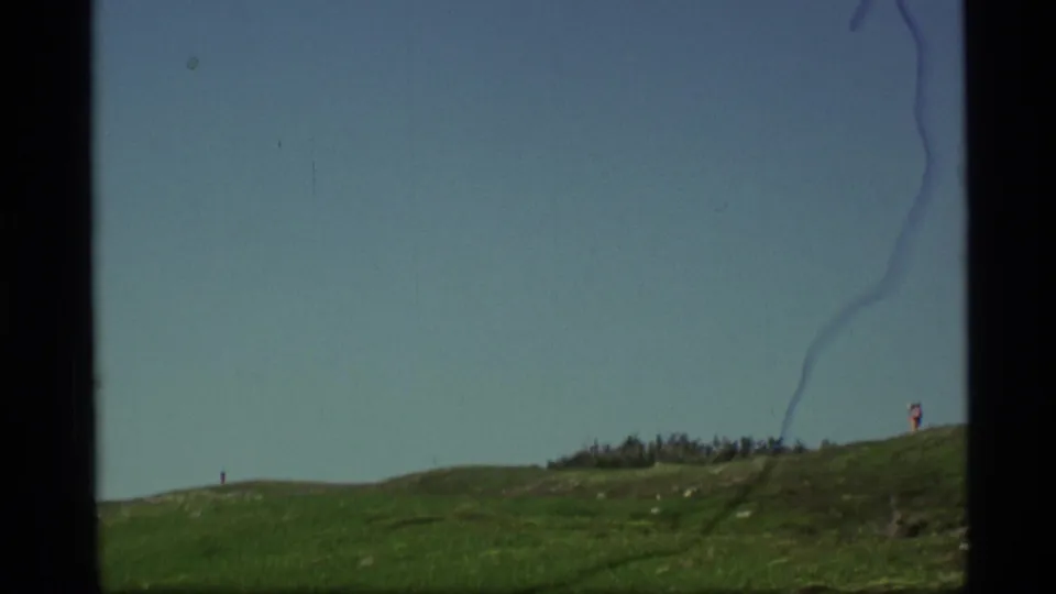 1977 Alaska home-movie frame with a green ridge, small trees, clear sky, and a tiny distant hiker at the right edge.