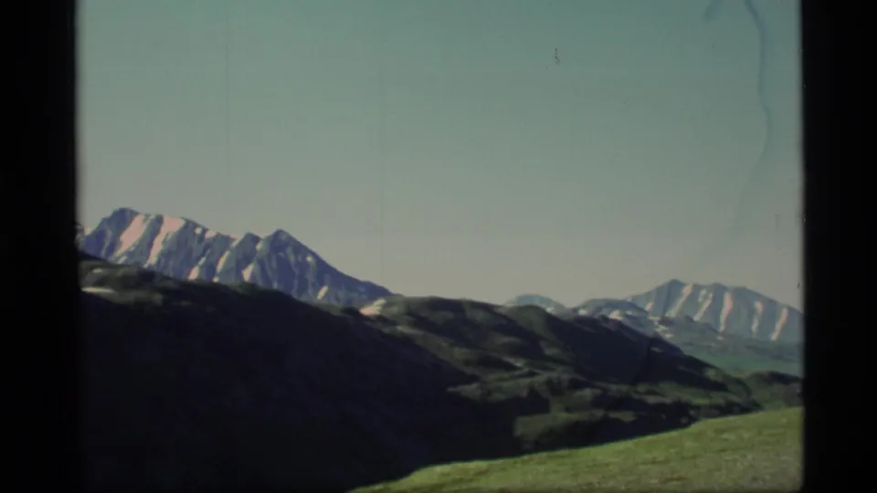 1977 Alaska 8mm landscape frame showing snow-streaked mountains beyond dark foreground hills and open grass.