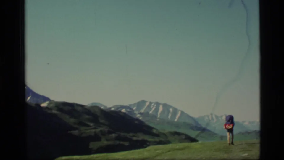 1977 Alaska 8mm frame showing a backpacker on a grassy hillside with a distant snow-streaked mountain range.