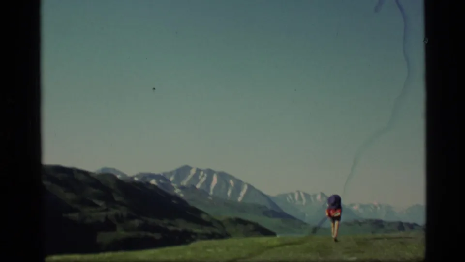 A lone backpacker crosses an alpine ridge in Alaska, with layered snow-capped peaks under a clear sky in 1977 8mm footage.