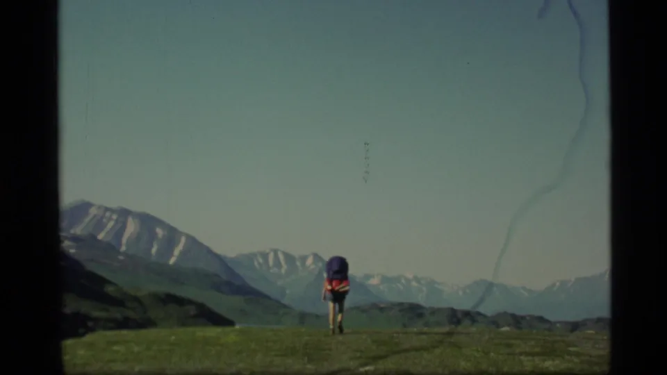 The hiker appears farther along the Alaska ridge, framed by high mountain peaks and a wide blue sky in 1970s home-movie footage.